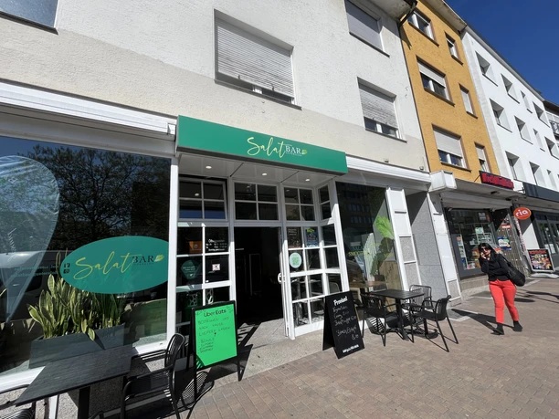 SalatBar Eingang der SalatBar mit grünem Schild, Sitzplätzen draußen und Schiefertafeln vor der Tür.