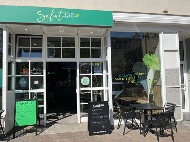 Eingang der SalatBar mit grünem Schild, Tischen im Außenbereich und Angebotstafel
