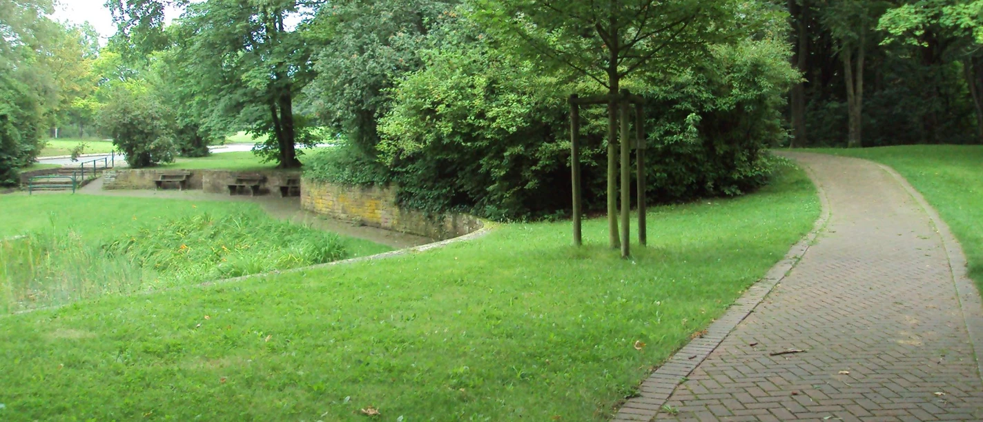 Gepflasterter Kurweg schlängelt sich durch eine grüne Parkanlage mit Bäumen und Sitzbänken.