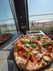 Pizza mit Parmaschinken und Rucola vor einem Fenster mit sonnigem Ausblick auf das Meer.