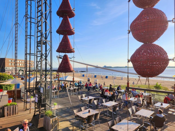 Panorama Weser-Strandbad 20240508_190439 (c) Dagmar Brandenburg_Erlebnis Bremerhaven_WEB.jpg Belebte Außenterrasse am Strandcafé Seelust mit roten Deko-Körben, Sandstrand und Wasser.