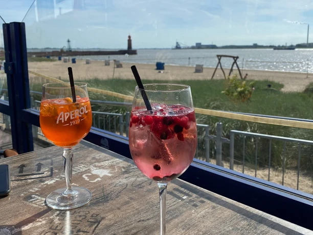 Seelust Getränke Seelust mit Ausblick Weser-Strandbad 2 (c) Mailin Knoke_Erlebnis Bremerhaven NICHT VERWENDEN!!!_WEB.jpg Zwei bunte Cocktails auf einem Tisch, dahinter Sandstrand, Meer und ein roter Leuchtturm.