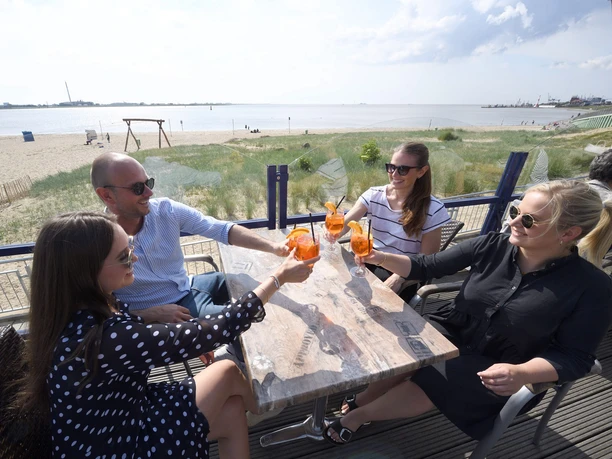 Seelust 10.6.20 Erlebnis3 (c) Wolfhard Scheer_Erlebnis Bremerhaven_WEB.jpg Vier Personen stoßen auf der sonnigen Terrasse am Sandstrand fröhlich mit ihren Drinks an.