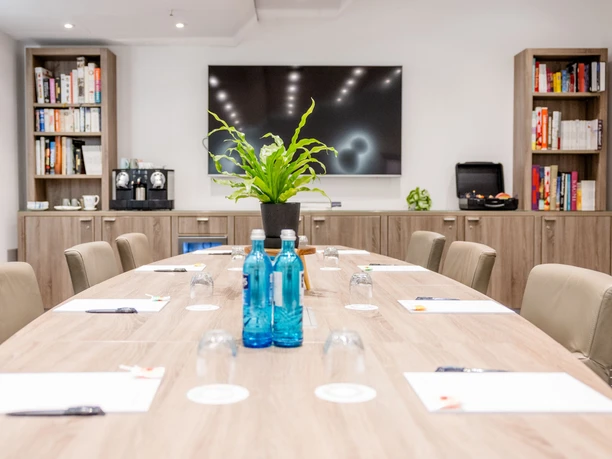 Motel One Frankfurt-Hauptbahnhof - Konferenzraum Moderner Konferenzraum mit einem langen Holztisch, blauen Wasserflaschen, Ledersesseln und einem großen Bildschirm im Hintergrund.
