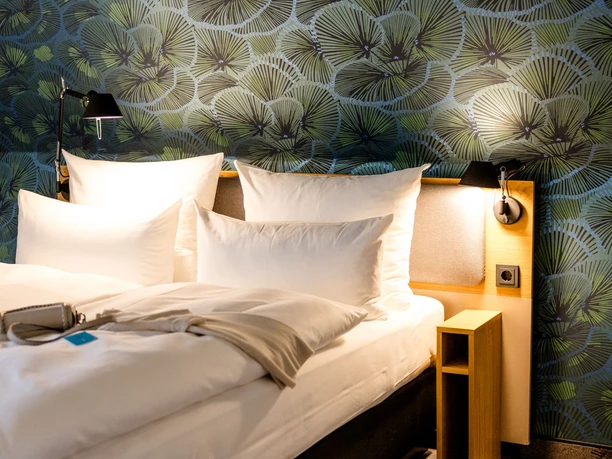 Motel One Frankfurt-Hauptbahnhof - Hotelzimmer Gemachtes Hotelbett mit weißen Kissen vor einer grün gemusterten Tapete.