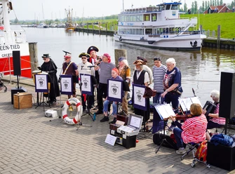 Sonntagskonzerte am Hafen Greetsiel Ein Shantychor in Piratenkostümen musiziert am Hafenbecken vor einem weißen Schiff.