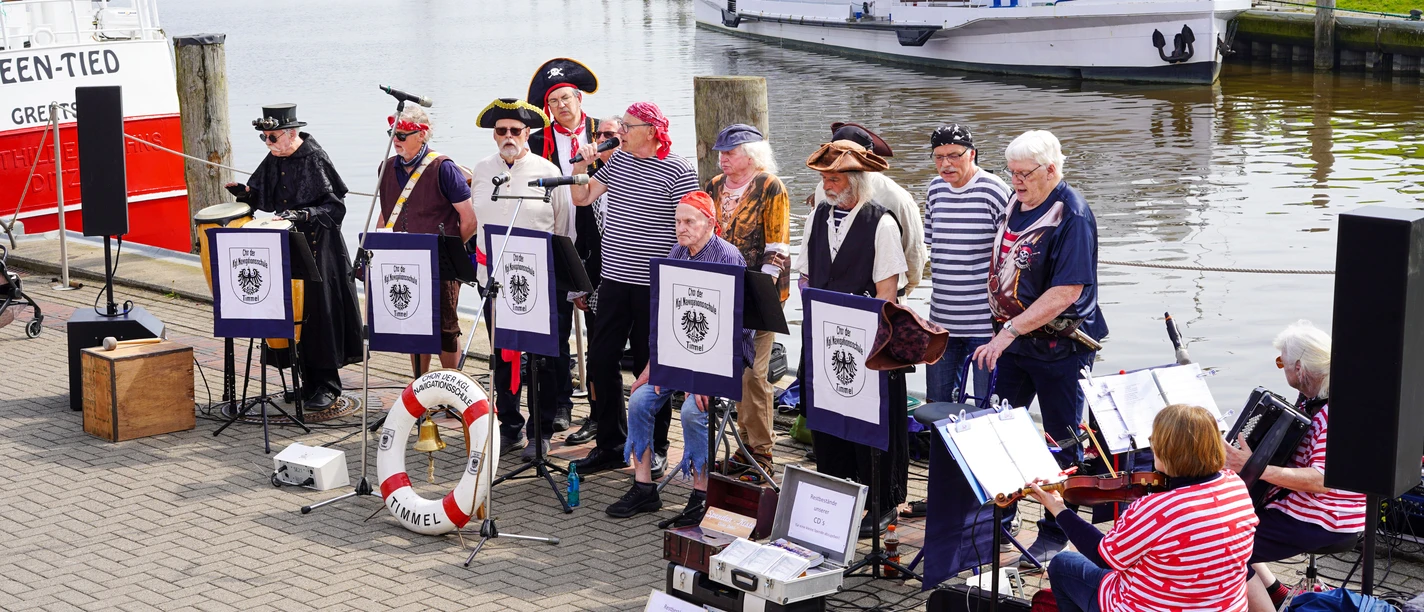 Sonntagskonzerte am Hafen Greetsiel Ein Shantychor in Piratenkostümen musiziert am Hafenbecken vor einem weißen Schiff.