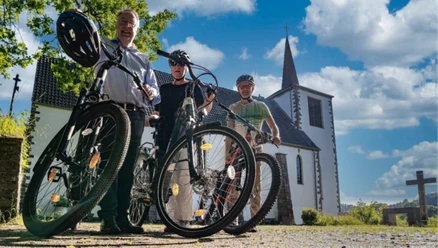 Radpilgern Drei Radfahrende mit Fahrrädern vor einer kleinen Kirche unter blauem Himmel