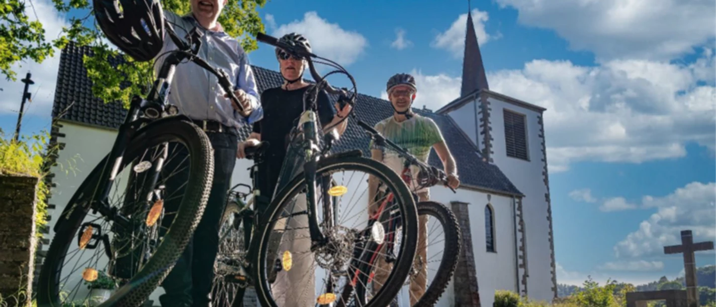 Radpilgern Drei Radfahrende mit Fahrrädern vor einer kleinen Kirche unter blauem Himmel
