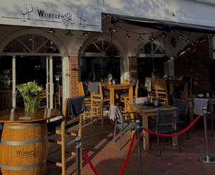 weinbar-aussenansicht.jpg Außenbereich einer Weinbar mit bestuhlter Terrasse, Rundbogenfenstern und Weinfass.