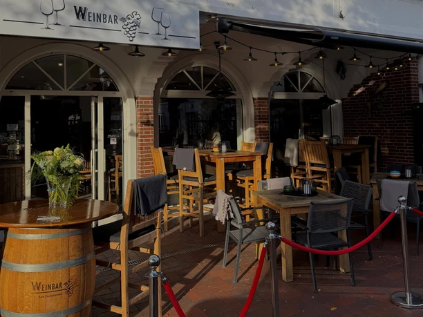 weinbar-aussenansicht.jpg Außenbereich einer Weinbar mit bestuhlter Terrasse, Rundbogenfenstern und Weinfass.