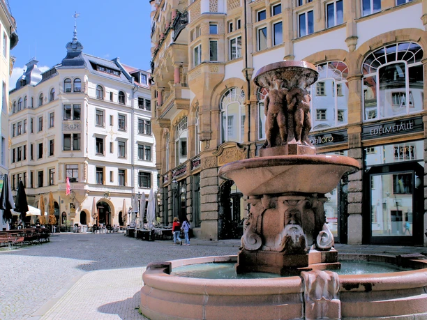 View of the Barfußgässchen with its row of bars and outdoor seating and the Lipsia fountain with its water feature in the foreground.