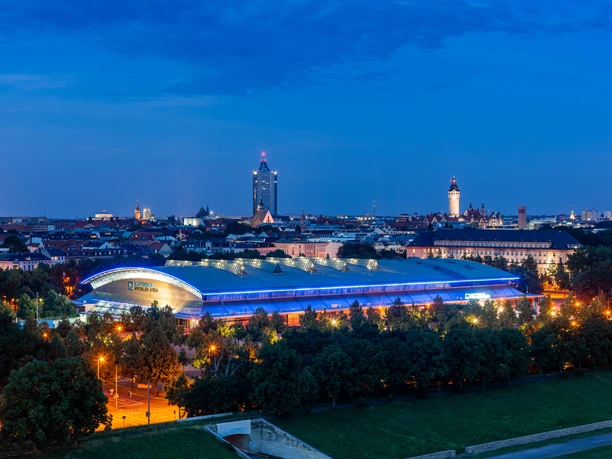 Widok na QUARTERBACK Immobilien Arena o zmierzchu z panoramą Lipska w tle, w tym City Tower i Nowy Ratusz.