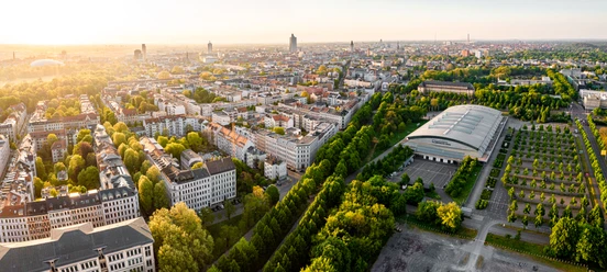 The aerial view shows the Waldstraßenviertel as the sun rises with the Leipzig skyline in the background and the QUARTERBACK Immobilien ARENA in the right-hand center of the picture.