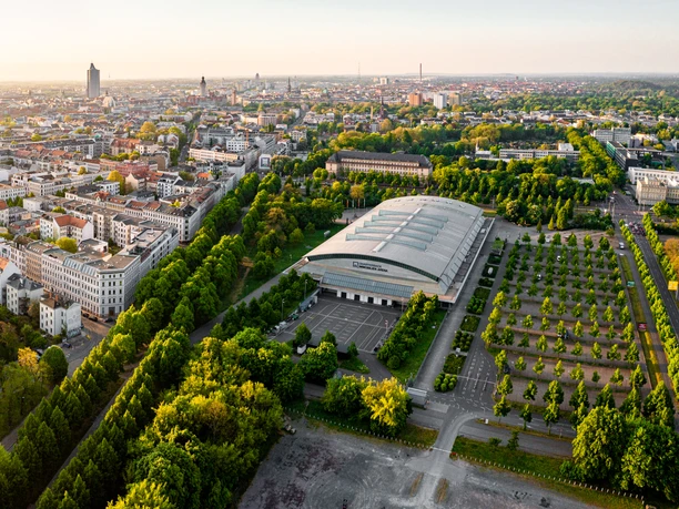 Widok z lotu ptaka przedstawia Waldstraßenviertel w świetle wschodzącego słońca z panoramą Lipska w tle i QUARTERBACK Immobilien ARENA w prawym środkowym rogu zdjęcia.