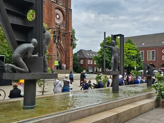 20240608_135806 (c) Tanja Mehl_Erlebnis Bremerhaven_WEB.jpg Werftbrunnen in Bremerhaven: Arbeiterskulpturen im Wasserbecken vor einer roten Backsteinkirche.