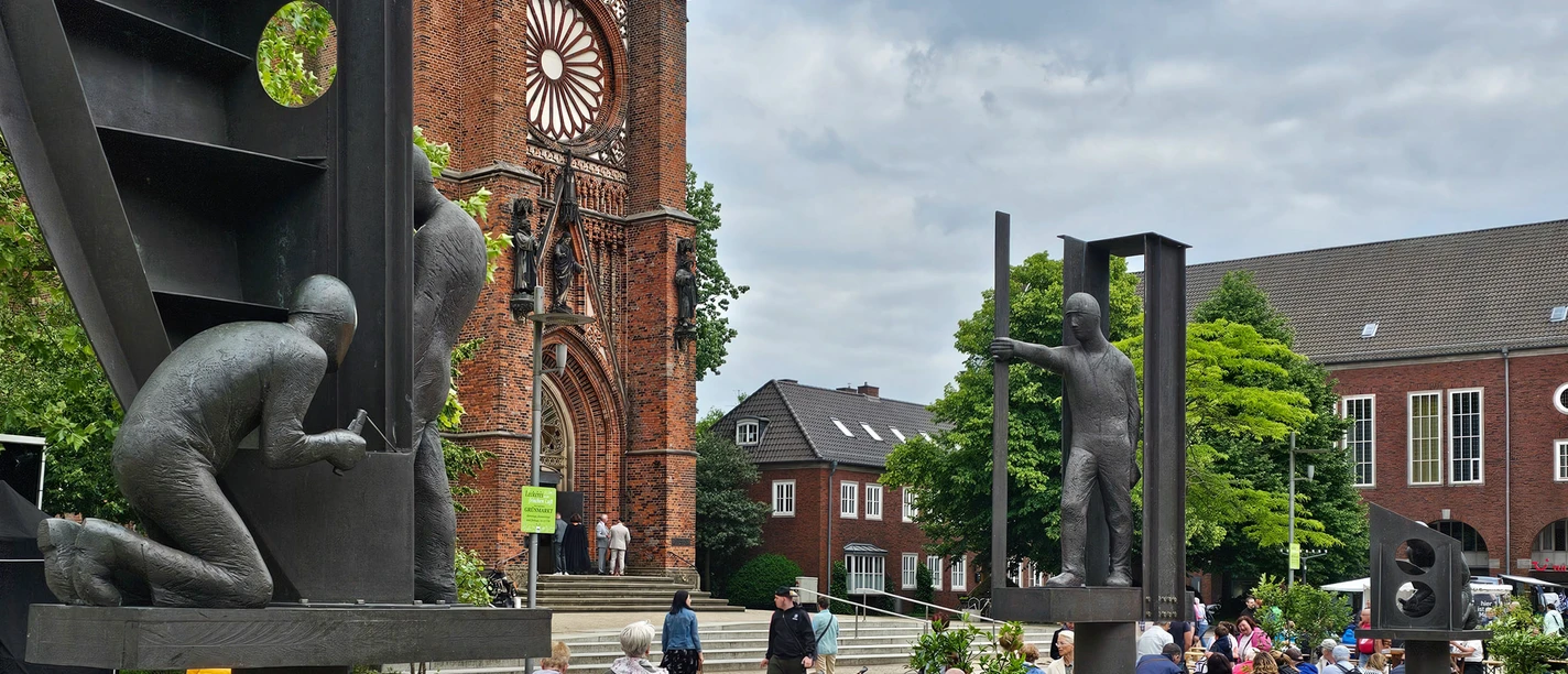 20240608_135806 (c) Tanja Mehl_Erlebnis Bremerhaven_WEB.jpg Werftbrunnen in Bremerhaven: Arbeiterskulpturen im Wasserbecken vor einer roten Backsteinkirche.