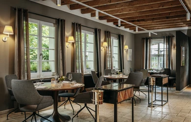 Gourmetrestaurant „Schote“ Eleganter Gastraum mit gedeckten Tischen, großen Fenstern, warmem Licht und Holzbalkendecke