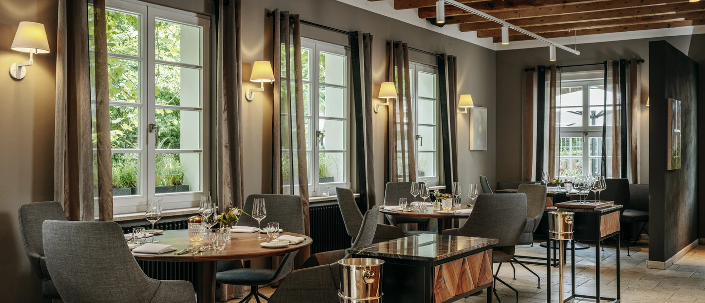 Gourmetrestaurant „Schote“ Eleganter Gastraum mit gedeckten Tischen, großen Fenstern, warmem Licht und Holzbalkendecke