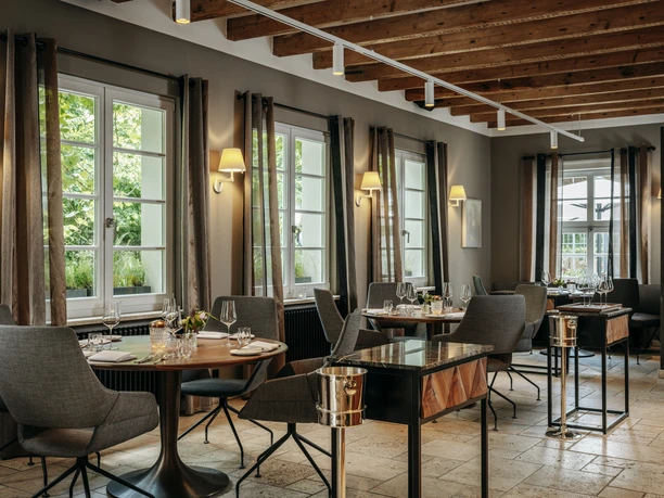 Gourmetrestaurant „Schote“ Eleganter Gastraum mit gedeckten Tischen, großen Fenstern, warmem Licht und Holzbalkendecke