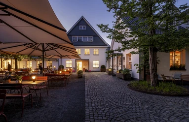 Gourmetrestaurant „Schote“ Beleuchteter Innenhof mit Tischen, großen Schirmen und historischem Gebäude bei Abenddämmerung
