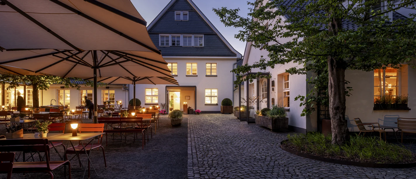 Gourmetrestaurant „Schote“ Beleuchteter Innenhof mit Tischen, großen Schirmen und historischem Gebäude bei Abenddämmerung