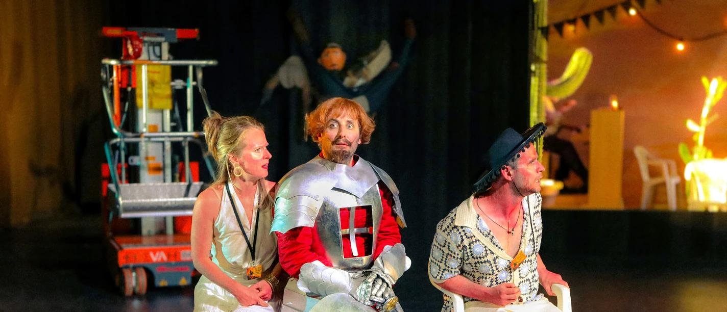 Probenfoto aus „¿Qué pasa en la mancha?“ mit Julia Schubert, Bastian Reiber und Sebastian Grünewald Drei Darsteller sitzen auf der Bühne, einer in Ritterrüstung, umgeben von Requisiten.