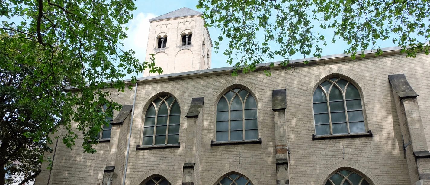 Kunststation St. Peter Helle Kirchenfassade mit spitzbogigen Fenstern und Turm, teils von Baumkronen beschattet