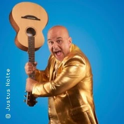 Ein Mann im goldenen Anzug hält eine Gitarre vor blauem Hintergrund und wirkt energiegeladen.