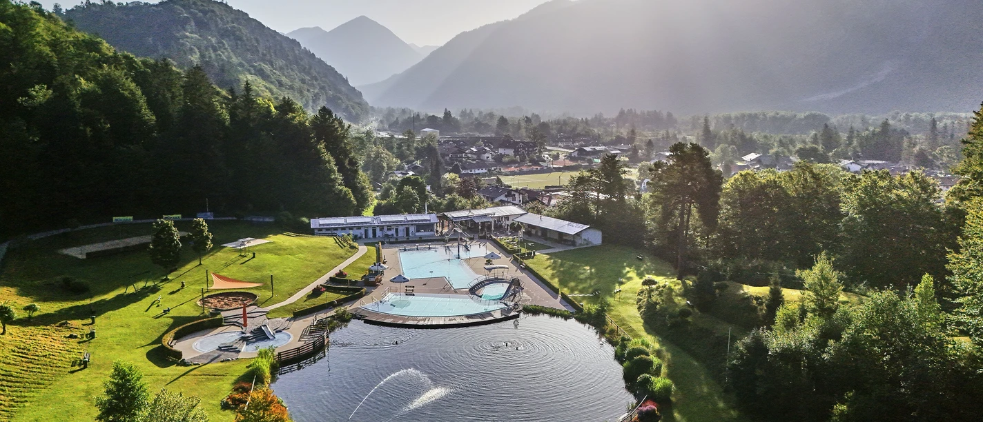 Alpenschwimmbad Oberau Freibad mit Becken und Naturbadesee in grüner Landschaft vor hoch aufragenden Bergen im Sonnenlicht