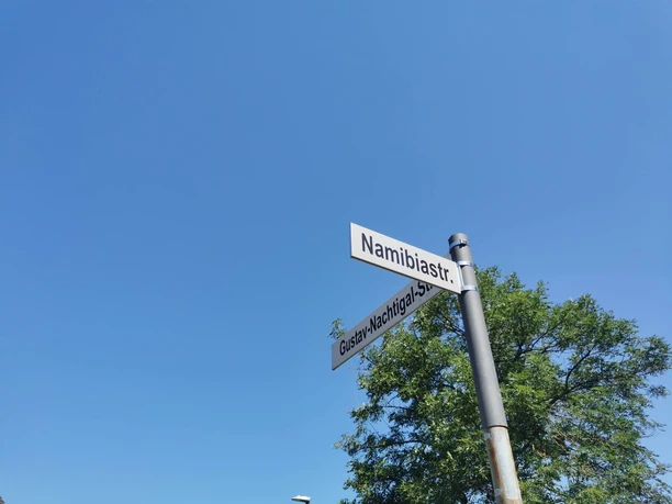 RollOut: On post-colonial traces Street signs Namibiastr. and Gustav-Nachtigal-Str. in front of a blue sky and a tree in the background