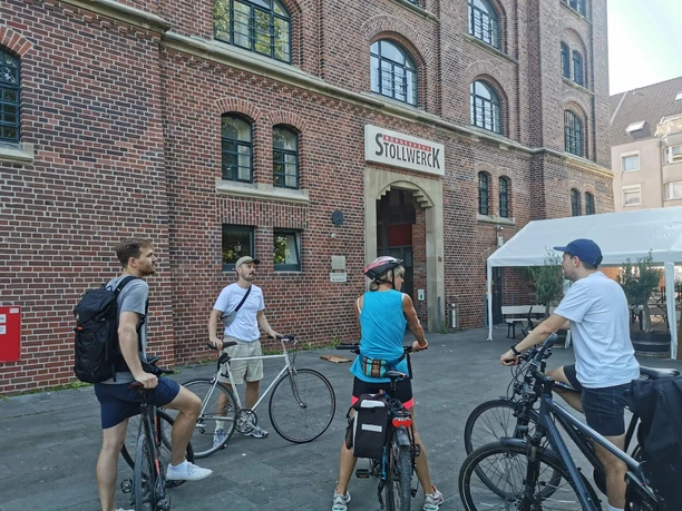 RollOut: Auf postkolonialen Spuren Radfahrende stehen vor einem roten Backsteingebäude mit dem Eingang zum Bürgerhaus Stollwerck