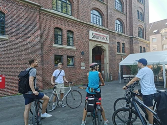 RollOut: On post-colonial traces Cyclists stand in front of a red brick building with the entrance to the Stollwerck community center