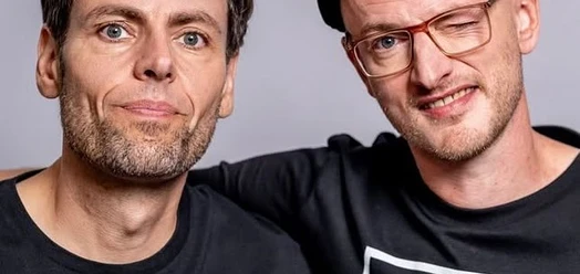 Porträt von Simon & Jan, zwei Männer in schwarzen Shirts vor grauem Hintergrund