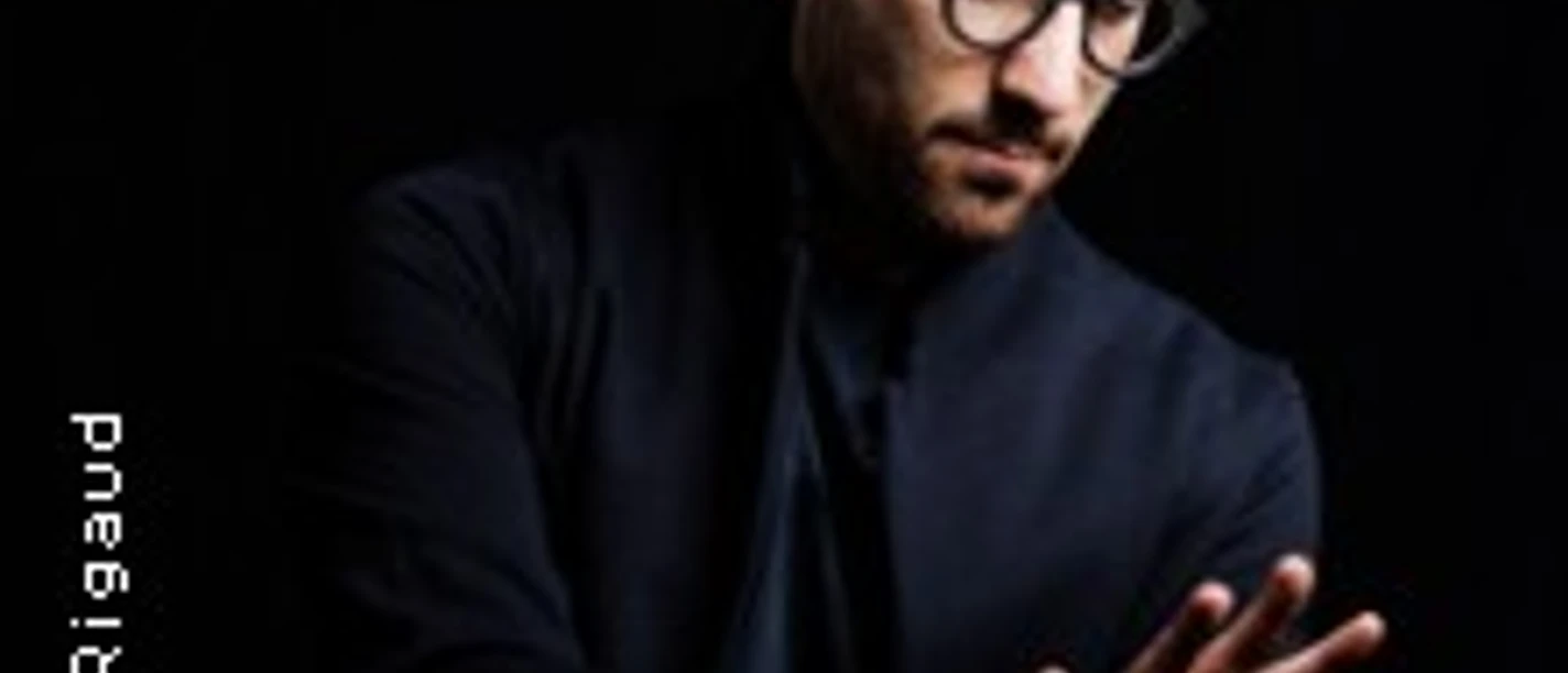 Mann mit Brille in dunkler Kleidung, der vor schwarzem Hintergrund beide Hände ausstreckt.