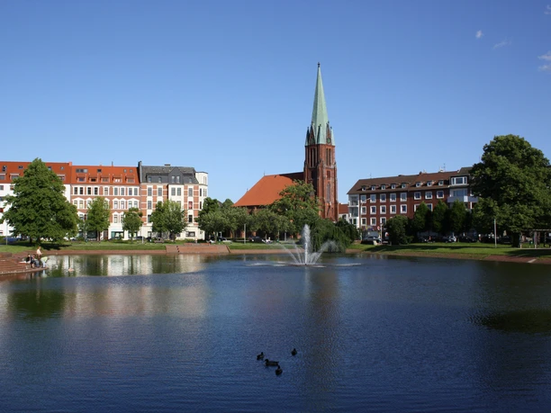 Springbrunnen am Holzhafen (c)Jens_Wöhlken__Holzhafen mit Christuskirche_1_WEB.jpg Springbrunnen im Holzhafen vor der roten Christuskirche unter blauem Himmel.