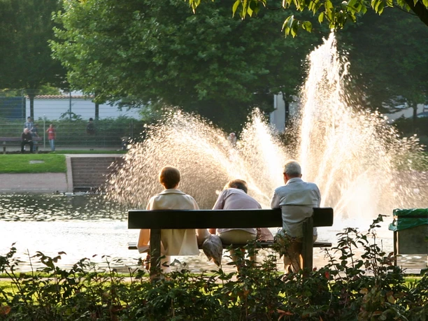 Springbrunnen am Holzhafen IMG_0834_WEB (c) Helmut Gross.jpg Drei Menschen auf einer Bank betrachten einen sprühenden Springbrunnen im Wasser.