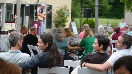 SE Saitenklänge Robert Carl Blank Bild Robert Scheuermeyer 260625 (21).JPG Open-Air‑Konzert mit Gitarrist auf kleiner Bühne und entspanntem Publikum an Tischen.