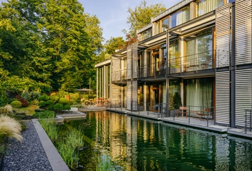 Nelson Müller Diepeschrather Mühle Moderner Bau mit großen Glasfronten am Wasser, umgeben von dichtem Grün.
