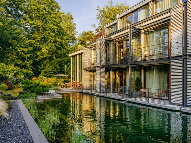 Nelson Müller Diepeschrather Mühle Moderner Bau mit großen Glasfronten am Wasser, umgeben von dichtem Grün.