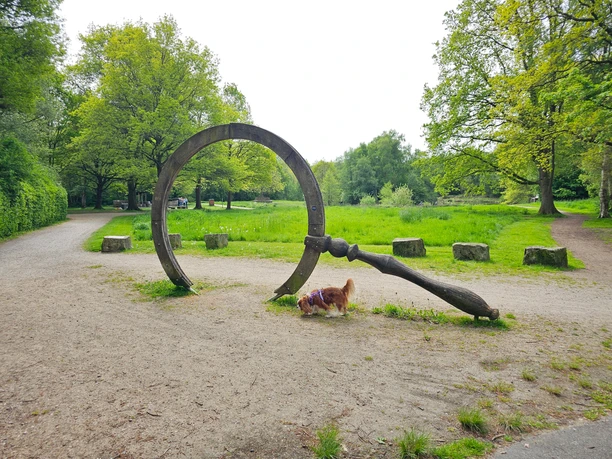 Erikasee 20260510_114053 (c) Tanja Mehl_Erlebnis Bremerhaven_bearb.jpg Riesige hölzerne Lupe auf einem Parkweg, darunter schnüffelt ein kleiner Hund.