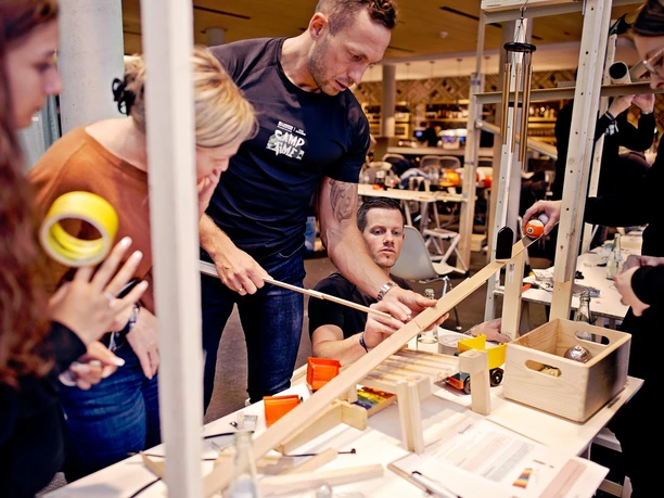 Impulsreaktion - 2 Teammitglieder arbeiten gemeinsam an einer komplexen Kettenreaktion mit Holzschienen und Kugeln auf einem Tisch.