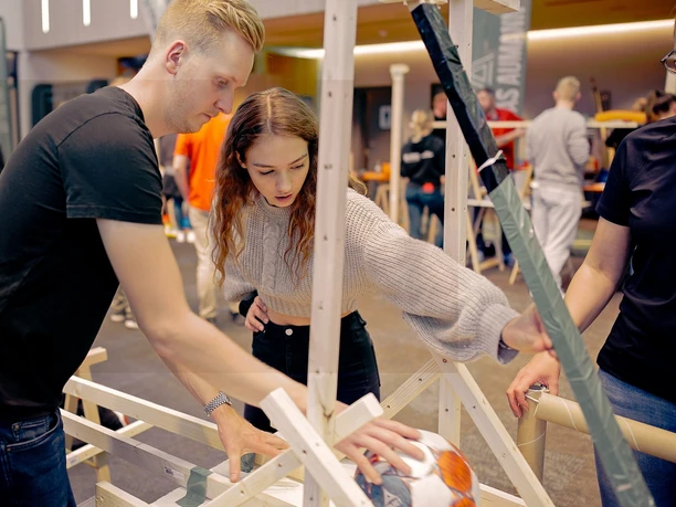 Impulsreaktion - 3 Drei Personen konzentrieren sich beim Teambuilding auf den Einbau eines Fußballs in eine hölzerne Kettenreaktions-Konstruktion.