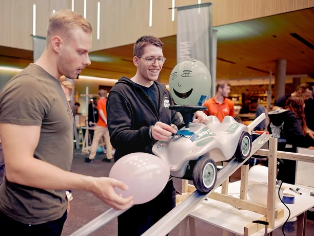 Impulsreaktion -1 Zwei Männer bauen bei einem Teamevent eine Kettenreaktion mit einem Bobby-Car auf einer Rampe und Luftballons auf.