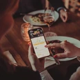 In einem Restaurant sitzt eine Gruppe von Personen an einem Tisch und isst, während eine Person einer anderen die Moveat-App auf ihrem Handy zeigt.