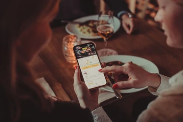 Kulinarischer Stadtrundgang_Moveat (c) Moveat.jpg In einem Restaurant sitzt eine Gruppe von Personen an einem Tisch und isst, während eine Person einer anderen die Moveat-App auf ihrem Handy zeigt.