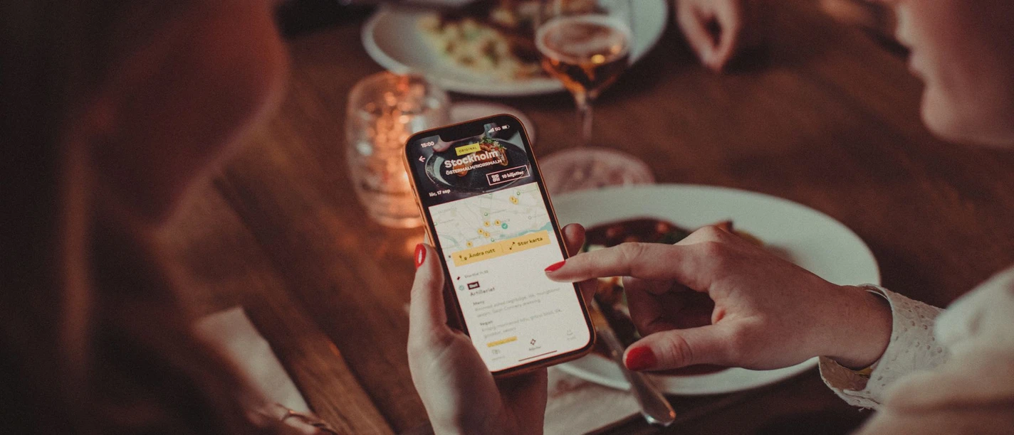 Kulinarischer Stadtrundgang_Moveat (c) Moveat.jpg In einem Restaurant sitzt eine Gruppe von Personen an einem Tisch und isst, während eine Person einer anderen die Moveat-App auf ihrem Handy zeigt.