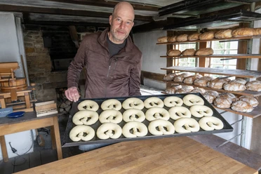 Museumsbäcker Stephan Klug Mann hält Blech mit rohen Teiglingen in historischer Backstube mit frischem Brot im Regal.