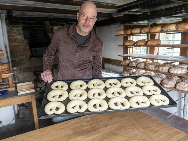 Museumsbäcker Stephan Klug Mann hält Blech mit rohen Teiglingen in historischer Backstube mit frischem Brot im Regal.