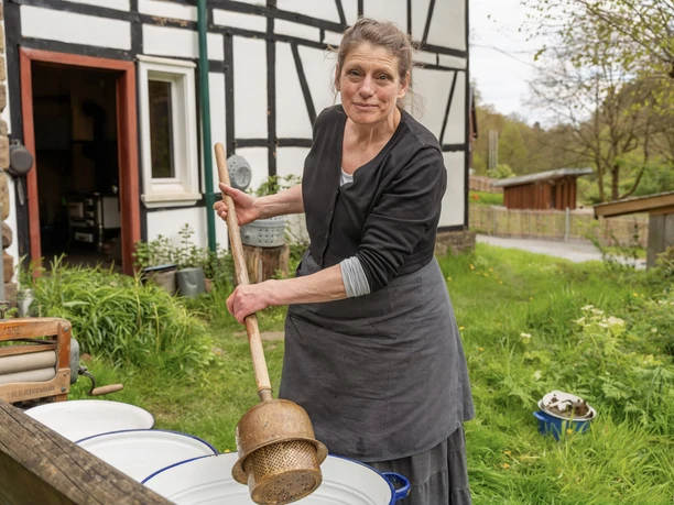Hauswirtschafterin Elisabeth Walter Frau rührt mit langem Holzstab in Waschzubern vor einem Fachwerkhaus in grüner Umgebung.